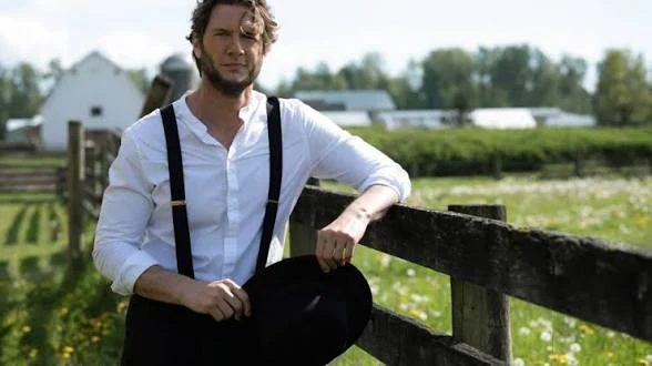 Ryan McPartlin holding a black hat beside a wooden fence in the countryside